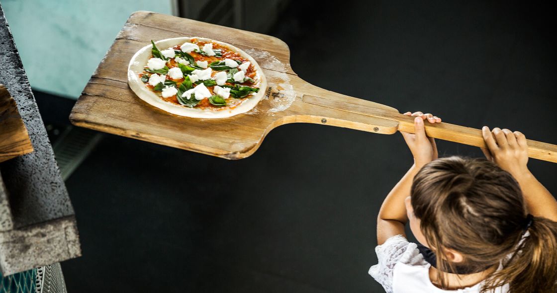 A Girl Holding A Wooden Paddle Over A Pizza On A Wooden Board