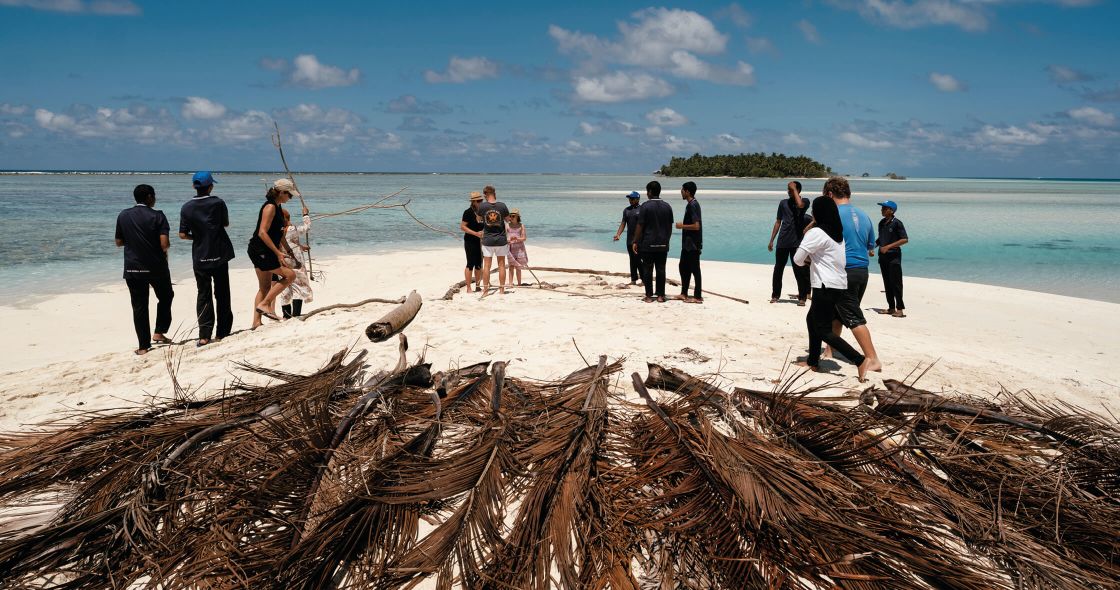 A Group Of People Standing On A Beach With A Large Pile Of Wood