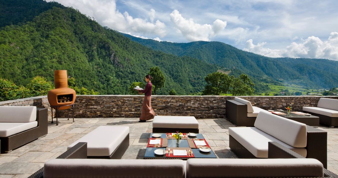 A Person Standing On A Patio With A Table And Chairs And A Mountain In The Background
