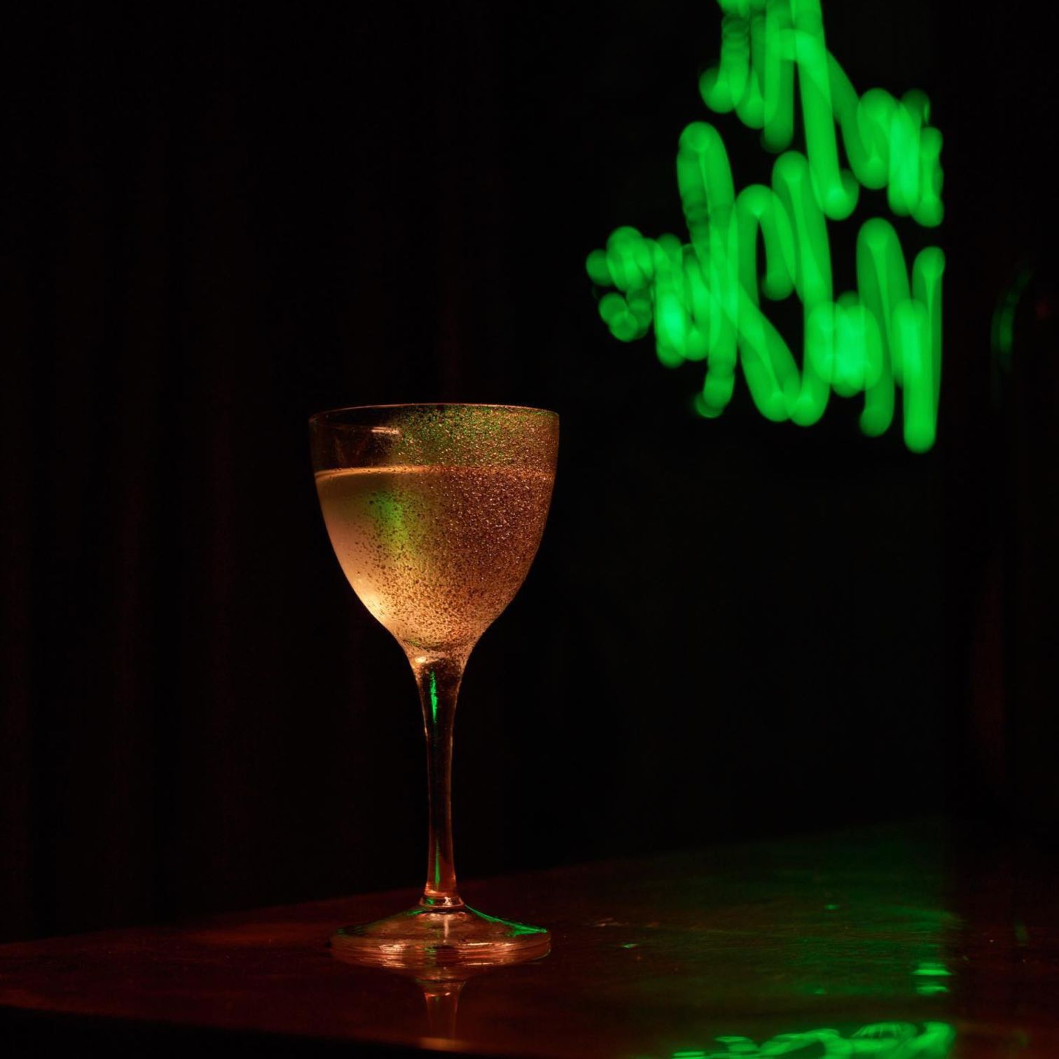 A Glass Of Wine With A Green Neon Sign In The Background